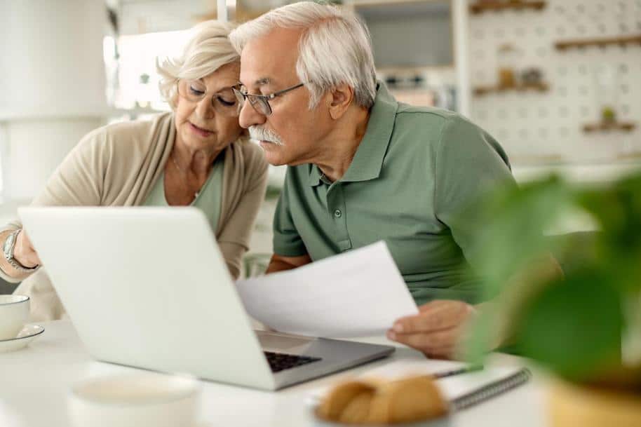 Elderly couple reviewing paperwork in front of a laptop on a table.