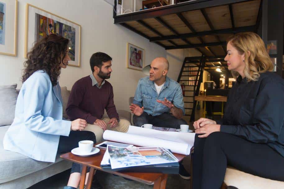 A group of people having a conversation while sitting around a coffee table with blueprints on it.