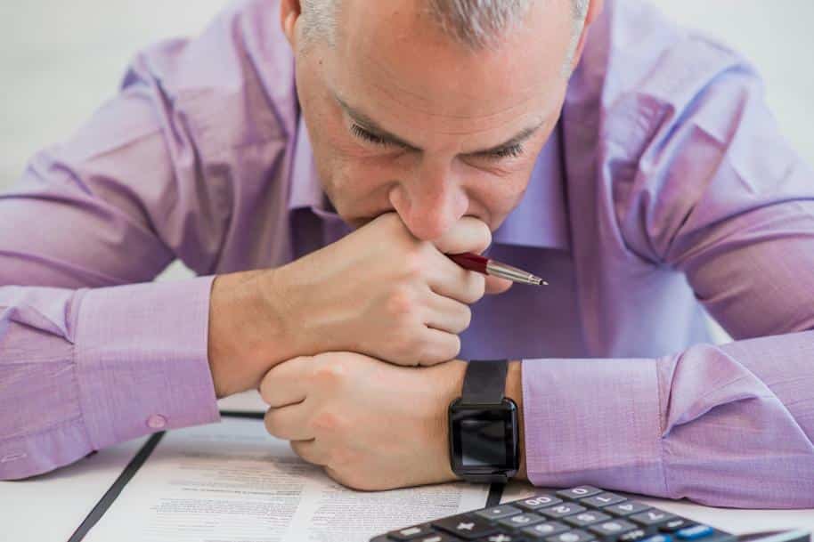 Closeup on a businessman looking sad, resting his face against his crossed fists, holding a pen and looking at a paper form.
